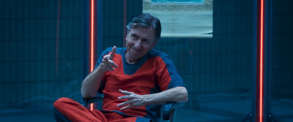 エレガントな囚人服を着て椅子に座り、ガラスの容器に座っているティム・ロス。